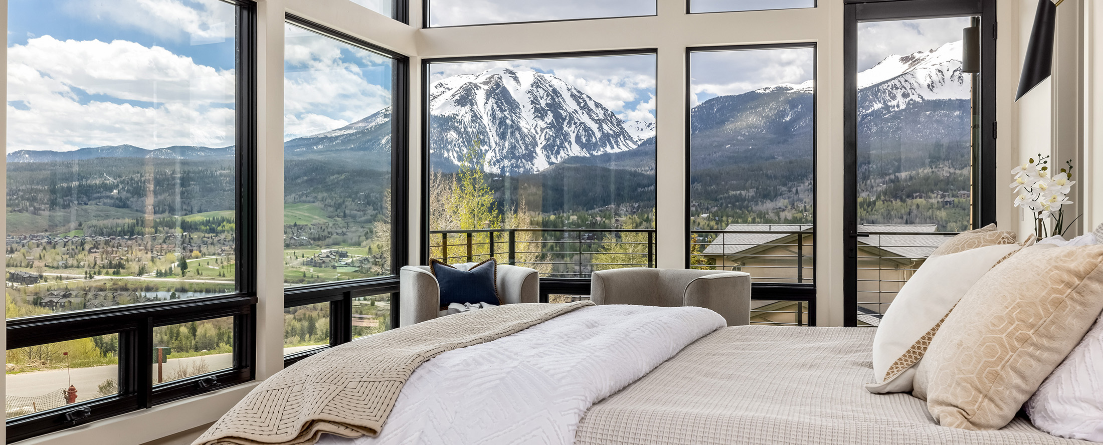 Luxury custom home bedroom with floor-to-ceiling corner windows and panoramic snowcapped mountain views, styled with modern neutral bedding