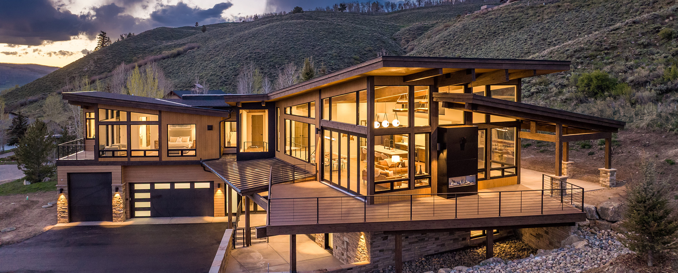 Modern mountain custom home at dusk with expansive glass walls, cantilevered deck railings, and warm interior lighting on a hillside lot