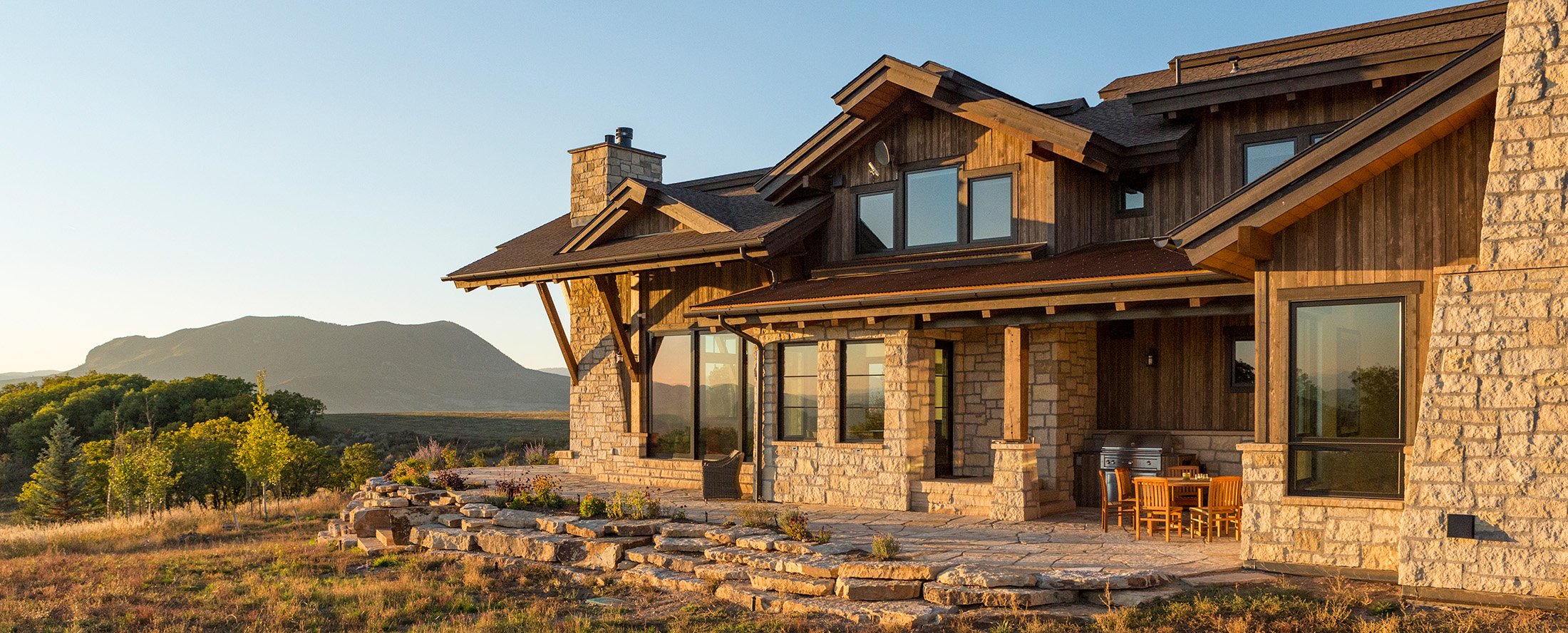 Rustic luxury custom home exterior with stone-and-wood siding, covered patio, and mountain views at sunset