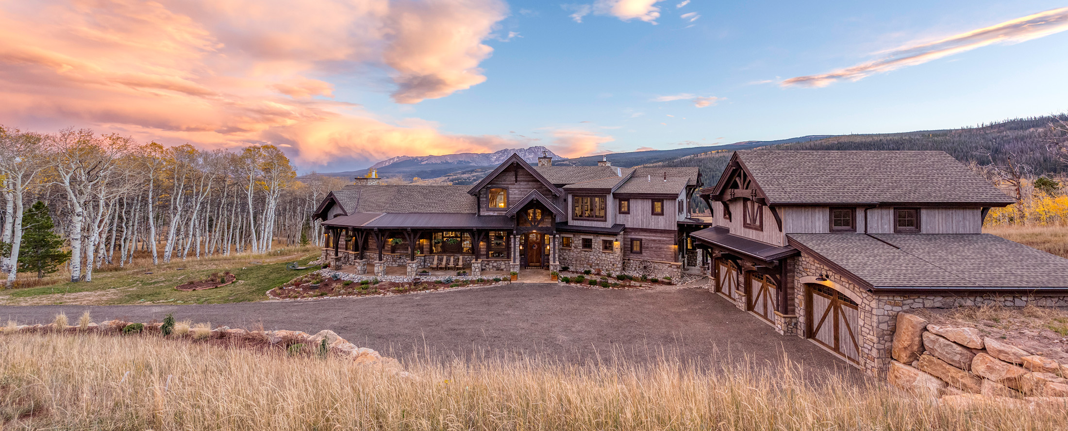 Rustic luxury ranch-style custom home with stone and wood exterior, covered porch, and oversized garage at sunset in a mountain meadow setting