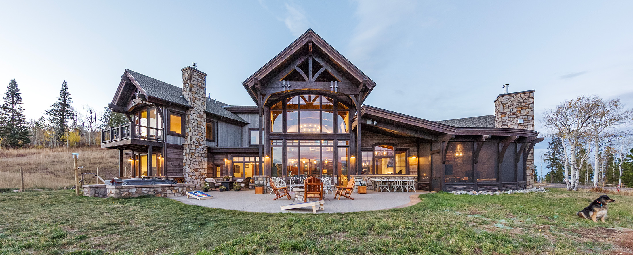 Luxury ranch-style custom home with timber trusses, stone chimneys, and expansive glass wall opening to an outdoor patio and entertaining area